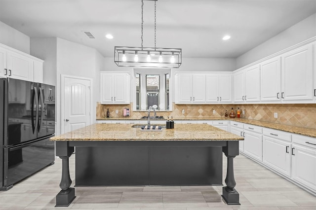 kitchen with a kitchen bar, black fridge, white cabinetry, light stone counters, and a kitchen island with sink