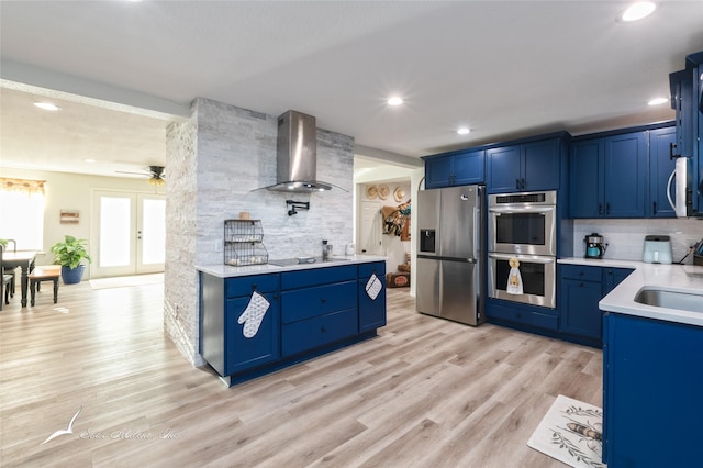 kitchen with blue cabinetry, appliances with stainless steel finishes, wall chimney range hood, light hardwood / wood-style floors, and backsplash