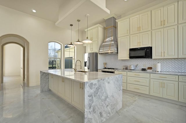 kitchen with cream cabinets, stainless steel fridge, hanging light fixtures, light stone countertops, and a center island with sink