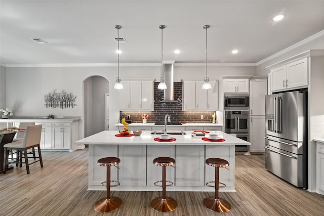 kitchen featuring white cabinets, hanging light fixtures, a kitchen island with sink, stainless steel appliances, and wall chimney range hood