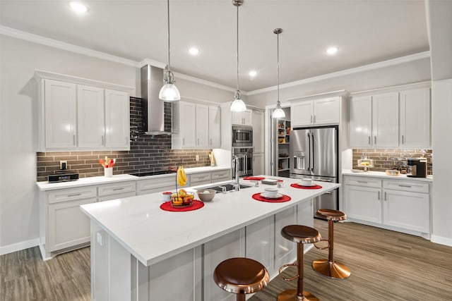 kitchen with decorative light fixtures, appliances with stainless steel finishes, wall chimney range hood, a kitchen island with sink, and white cabinets