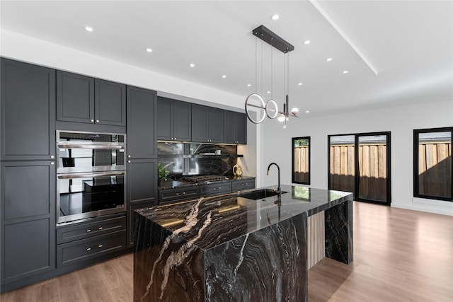 kitchen with sink, hanging light fixtures, appliances with stainless steel finishes, an island with sink, and dark stone counters