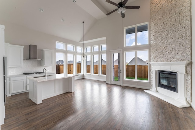 kitchen with a stone fireplace, sink, white cabinets, wall chimney range hood, and a center island with sink