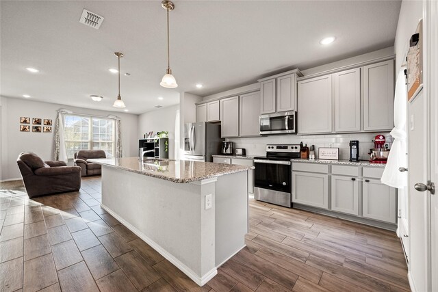 kitchen featuring gray cabinetry, a kitchen island with sink, stainless steel appliances, light stone counters, and decorative light fixtures