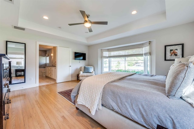 bedroom with ceiling fan, ensuite bath, a raised ceiling, and light hardwood / wood-style floors