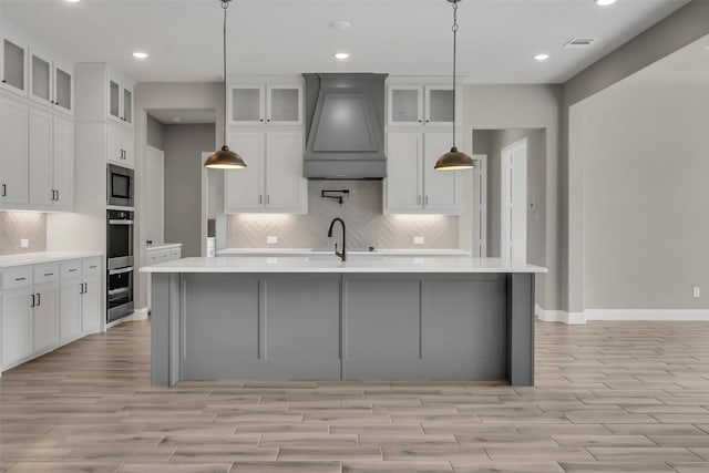 kitchen with pendant lighting, stainless steel microwave, decorative backsplash, custom exhaust hood, and a center island with sink