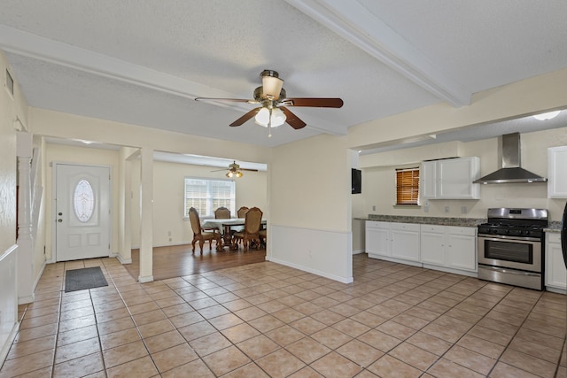 kitchen featuring light tile patterned flooring, stainless steel gas range, white cabinetry, beamed ceiling, and wall chimney range hood