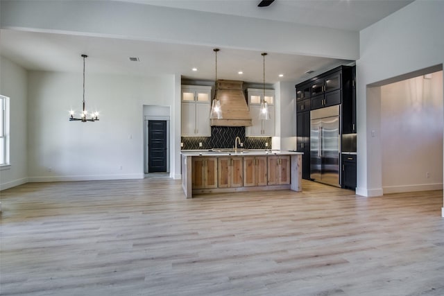 kitchen with tasteful backsplash, hanging light fixtures, stainless steel built in fridge, custom range hood, and a center island with sink