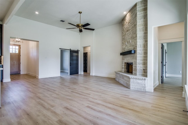 unfurnished living room with a barn door, ceiling fan with notable chandelier, a stone fireplace, and light hardwood / wood-style floors