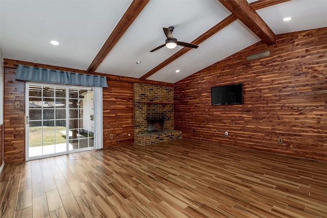 unfurnished living room with hardwood / wood-style flooring, ceiling fan, lofted ceiling with beams, a brick fireplace, and wood walls