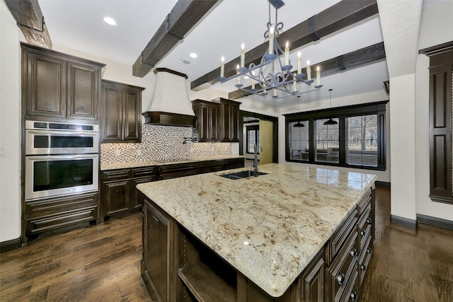 kitchen featuring sink, a kitchen island with sink, custom range hood, decorative backsplash, and stainless steel double oven