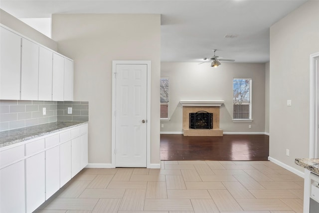 kitchen with ceiling fan, a fireplace, light stone countertops, white cabinets, and decorative backsplash