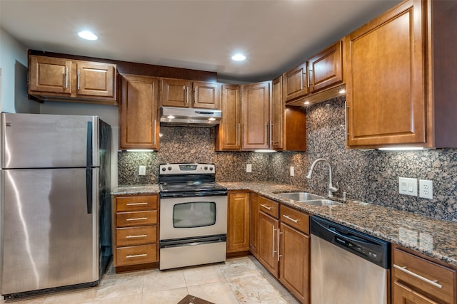 kitchen with dark stone countertops, under cabinet range hood, brown cabinetry, appliances with stainless steel finishes, and a sink