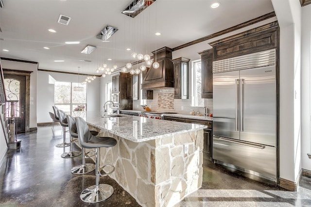 kitchen featuring a spacious island, light stone counters, built in refrigerator, decorative light fixtures, and custom range hood