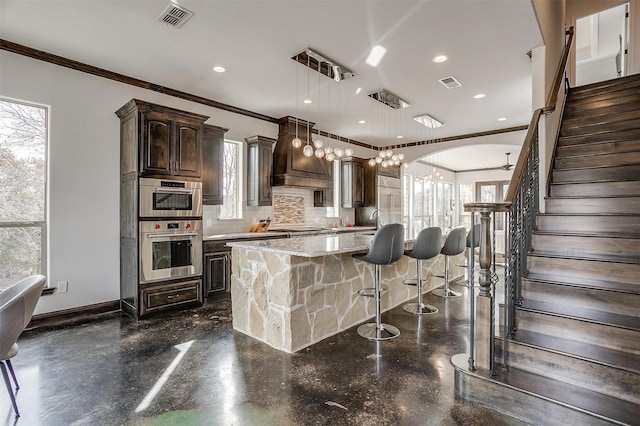 kitchen featuring pendant lighting, light stone counters, tasteful backsplash, dark brown cabinetry, and an island with sink