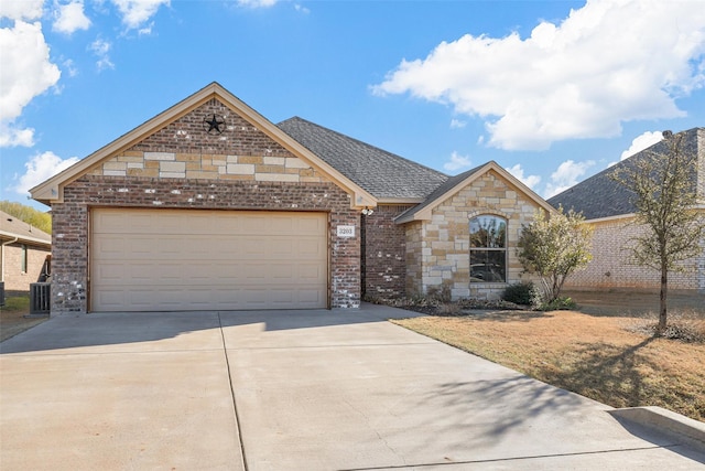 french country home with roof with shingles, concrete driveway, an attached garage, brick siding, and central AC unit