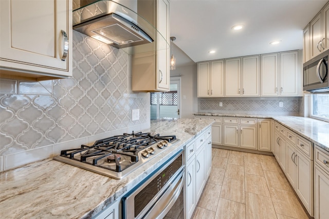 kitchen featuring tasteful backsplash, light stone counters, stainless steel appliances, wall chimney range hood, and cream cabinetry