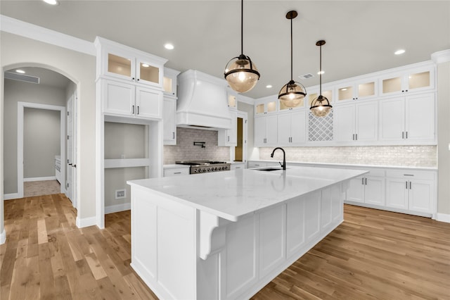 kitchen featuring pendant lighting, white cabinetry, light stone counters, a center island with sink, and custom exhaust hood