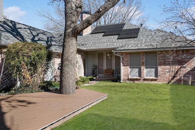 exterior space with brick siding, a shingled roof, roof mounted solar panels, a front lawn, and a chimney