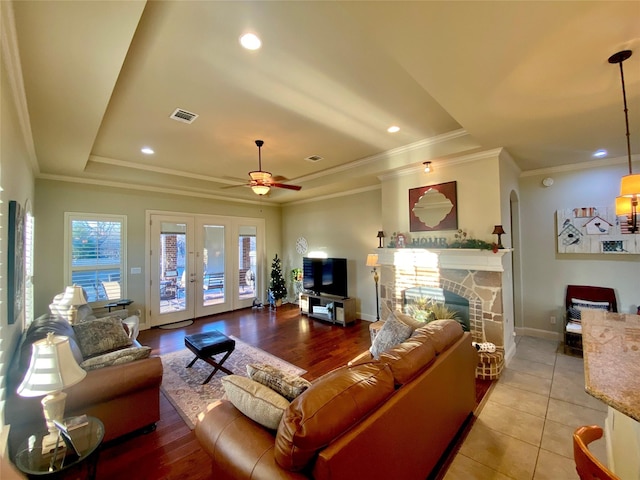 living room featuring french doors, a stone fireplace, ornamental molding, a tray ceiling, and ceiling fan