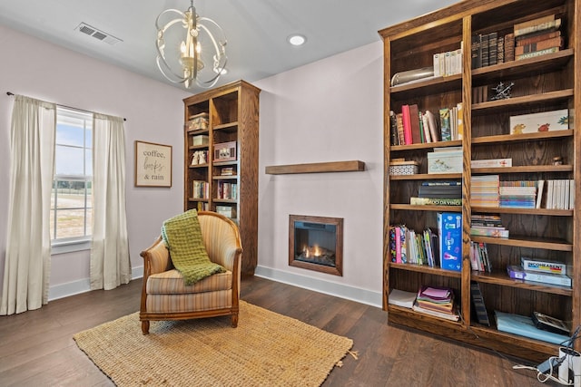 sitting room featuring a glass covered fireplace, visible vents, wood finished floors, baseboards, and a notable chandelier