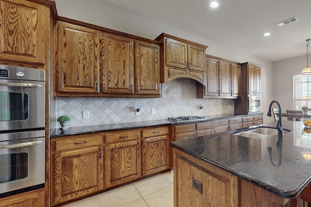 kitchen with sink, light tile patterned floors, dark stone counters, stainless steel appliances, and a kitchen island with sink