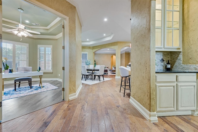 interior space featuring crown molding, ceiling fan, tasteful backsplash, a raised ceiling, and light wood-type flooring