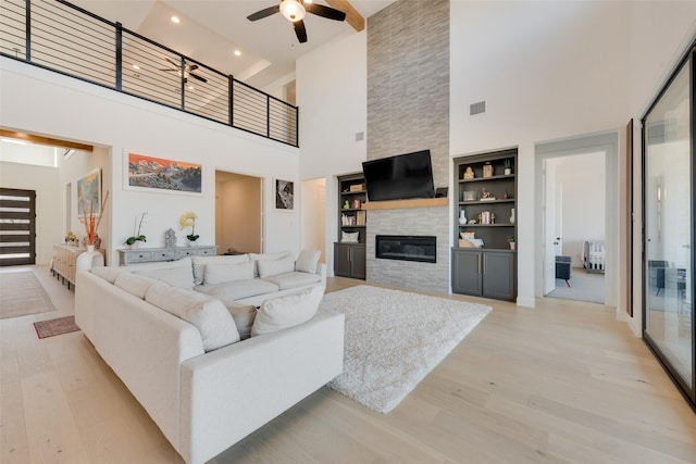 living room featuring a high ceiling, a tiled fireplace, ceiling fan, and light wood-style floors