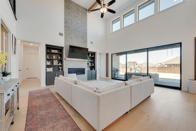 living area featuring a fireplace, built in shelves, light wood-style flooring, a ceiling fan, and a towering ceiling