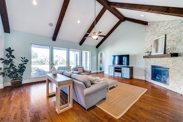 living room featuring dark wood-type flooring, ceiling fan, beam ceiling, high vaulted ceiling, and a stone fireplace