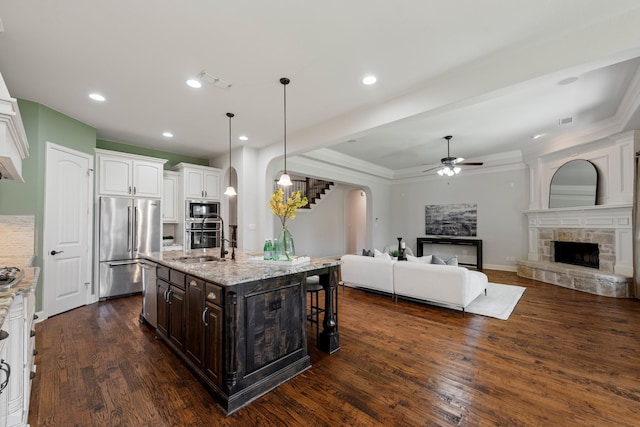 kitchen with arched walkways, dark brown cabinets, ceiling fan, a stone fireplace, and appliances with stainless steel finishes