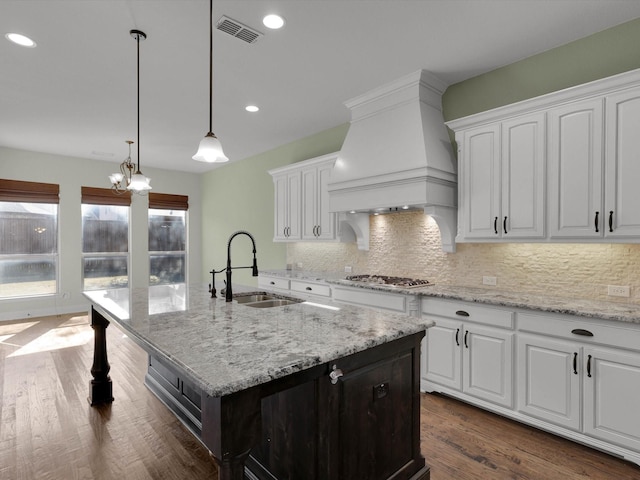 kitchen featuring sink, premium range hood, a kitchen island with sink, hanging light fixtures, and white cabinets