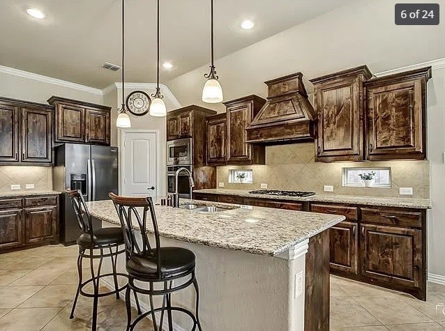 kitchen featuring an island with sink, appliances with stainless steel finishes, custom range hood, and decorative light fixtures