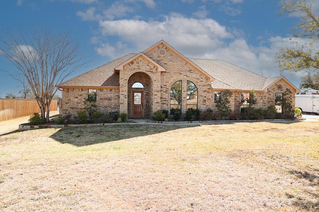 french country style house with brick siding, a shingled roof, a front yard, and fence