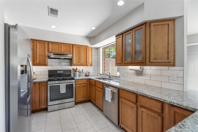 4808 Briarwood Ln, Fort Worth, TX 76103 - kitchen with stainless steel appliances, brown cabinetry, under cabinet range hood, light stone countertops, and decorative backsplash