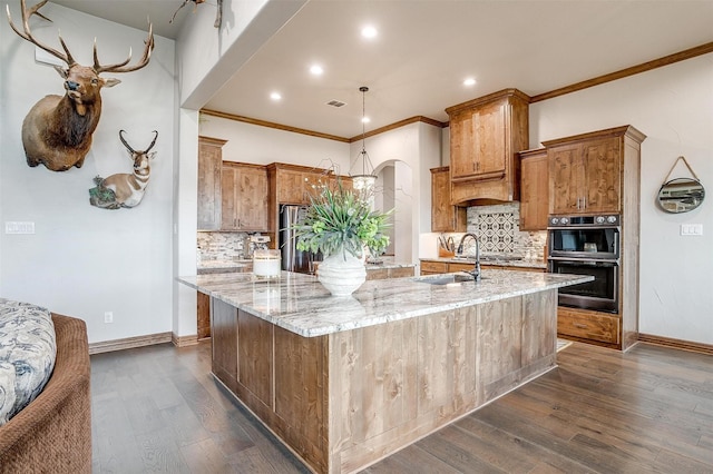 kitchen featuring crown molding, double wall oven, light stone countertops, dark hardwood / wood-style flooring, and a large island with sink