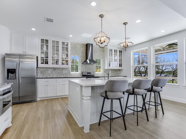 kitchen featuring appliances with stainless steel finishes, decorative light fixtures, white cabinets, wall chimney range hood, and a center island with sink