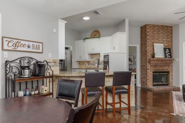 kitchen featuring stainless steel fridge, white cabinetry, backsplash, light stone countertops, and a brick fireplace