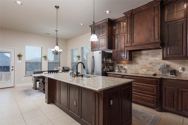 kitchen featuring dark brown cabinetry, sink, light stone counters, a center island with sink, and stainless steel refrigerator