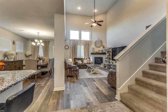 living room featuring a high ceiling, ceiling fan with notable chandelier, a fireplace, and dark hardwood / wood-style flooring