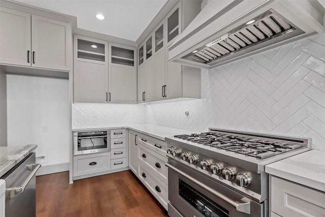 kitchen with dark wood finished floors, appliances with stainless steel finishes, glass insert cabinets, light stone countertops, and custom exhaust hood