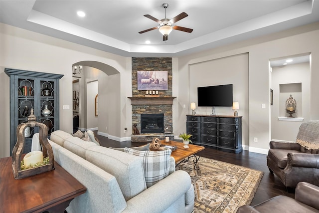 living room featuring dark hardwood / wood-style floors, a stone fireplace, a raised ceiling, and ceiling fan