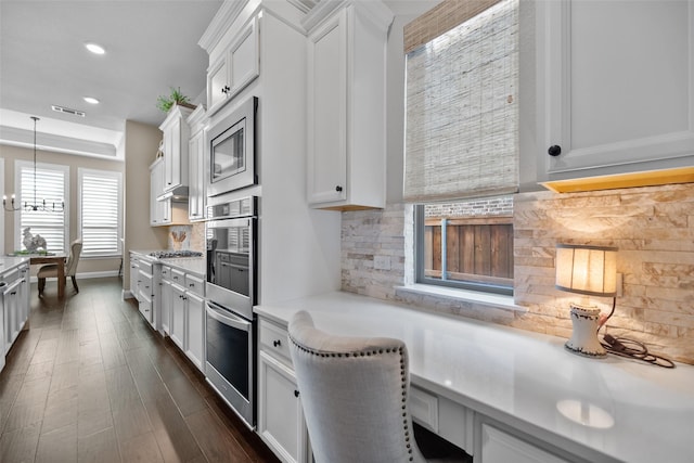 kitchen with white cabinetry, hanging light fixtures, backsplash, stainless steel appliances, and built in desk