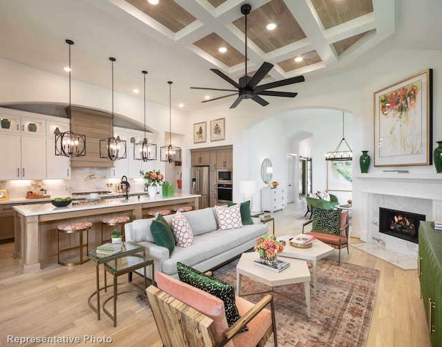 living room featuring a premium fireplace, a towering ceiling, coffered ceiling, light hardwood / wood-style floors, and ceiling fan with notable chandelier