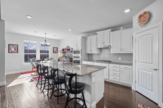 kitchen featuring appliances with stainless steel finishes, a center island with sink, light stone counters, white cabinets, and a kitchen bar