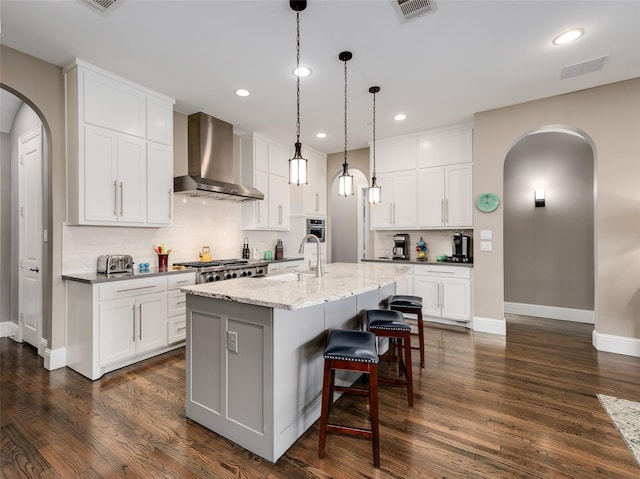 kitchen with wall chimney exhaust hood, light stone countertops, a center island with sink, and white cabinets