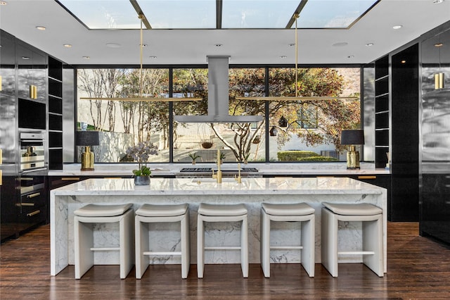 kitchen featuring a skylight, range hood, a breakfast bar area, light stone countertops, and dark cabinetry