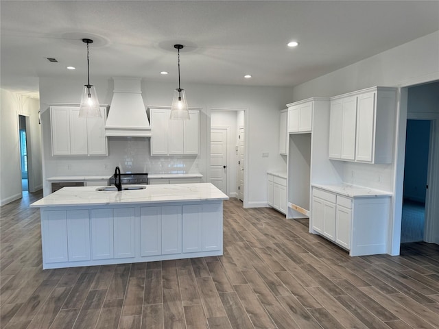 kitchen with sink, custom exhaust hood, white cabinetry, hanging light fixtures, and a kitchen island with sink