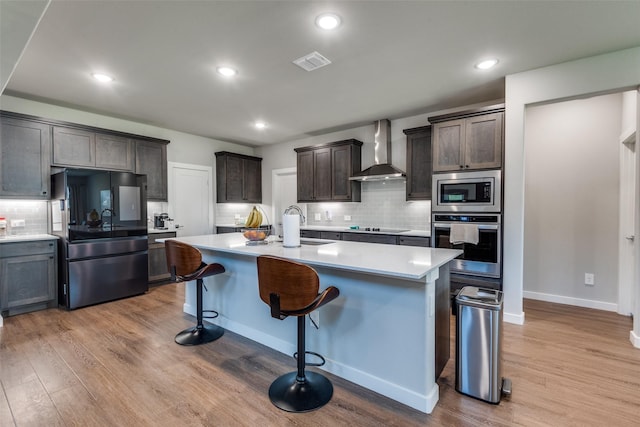 kitchen with dark brown cabinetry, light hardwood / wood-style flooring, stainless steel appliances, a kitchen island with sink, and wall chimney range hood