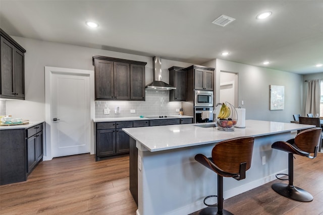 kitchen featuring dark brown cabinets, hardwood / wood-style floors, stainless steel appliances, a kitchen island with sink, and wall chimney range hood